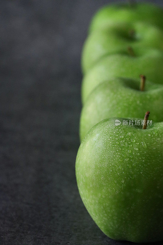 一排五个，绿色的史密斯苹果(Malus domestica)的特写图像，有茎，光滑的，有斑点的果皮，斑驳的深灰色背景，重点在前景，复制空间